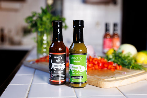 two bottles of hot sauce on a counter top with vegetables in the background